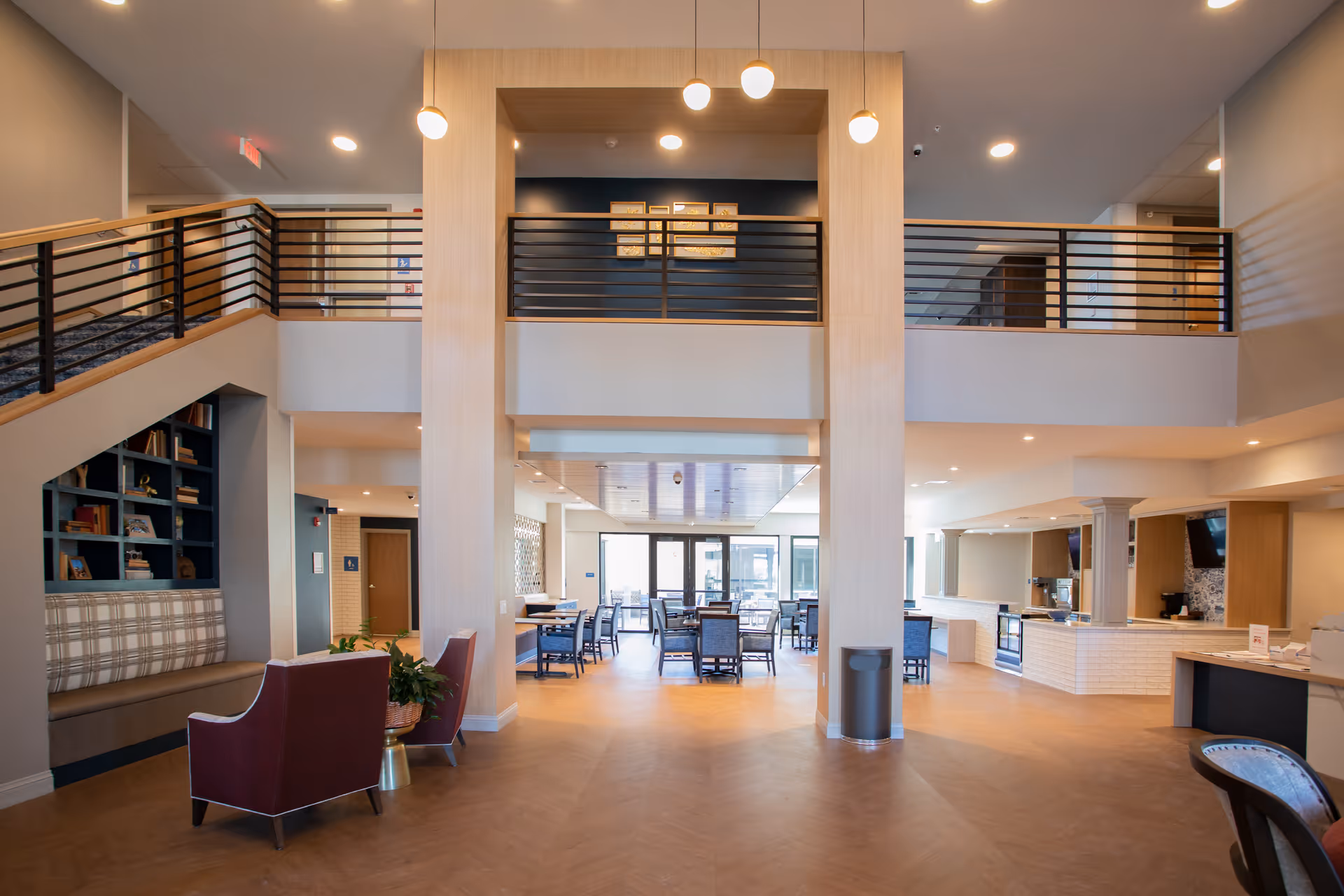 Spacious and modern interior of a senior living facility with a two-story ceiling, a staircase with black railings, seating areas with chairs and a built-in bench, and a dining area with tables and chairs in the background. The space is well-lit with ceiling lights and pendant lights, featuring light wood flooring and neutral-colored walls.