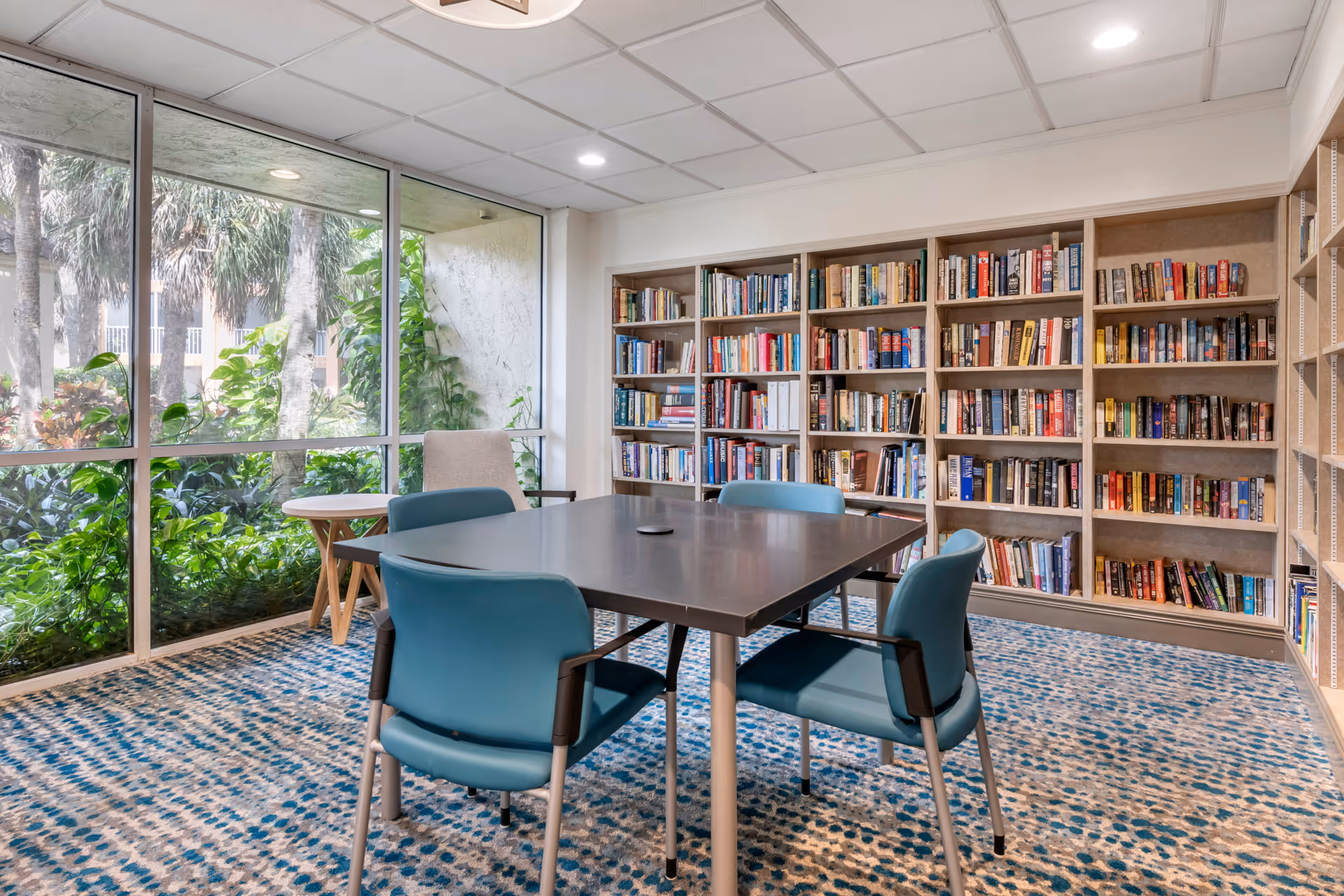 A bright room with large windows showing greenery outside. Inside, there is a square table surrounded by four blue chairs and one beige chair. Behind the table, there are bookshelves filled with books. The floor is covered with a patterned carpet in shades of blue and beige.