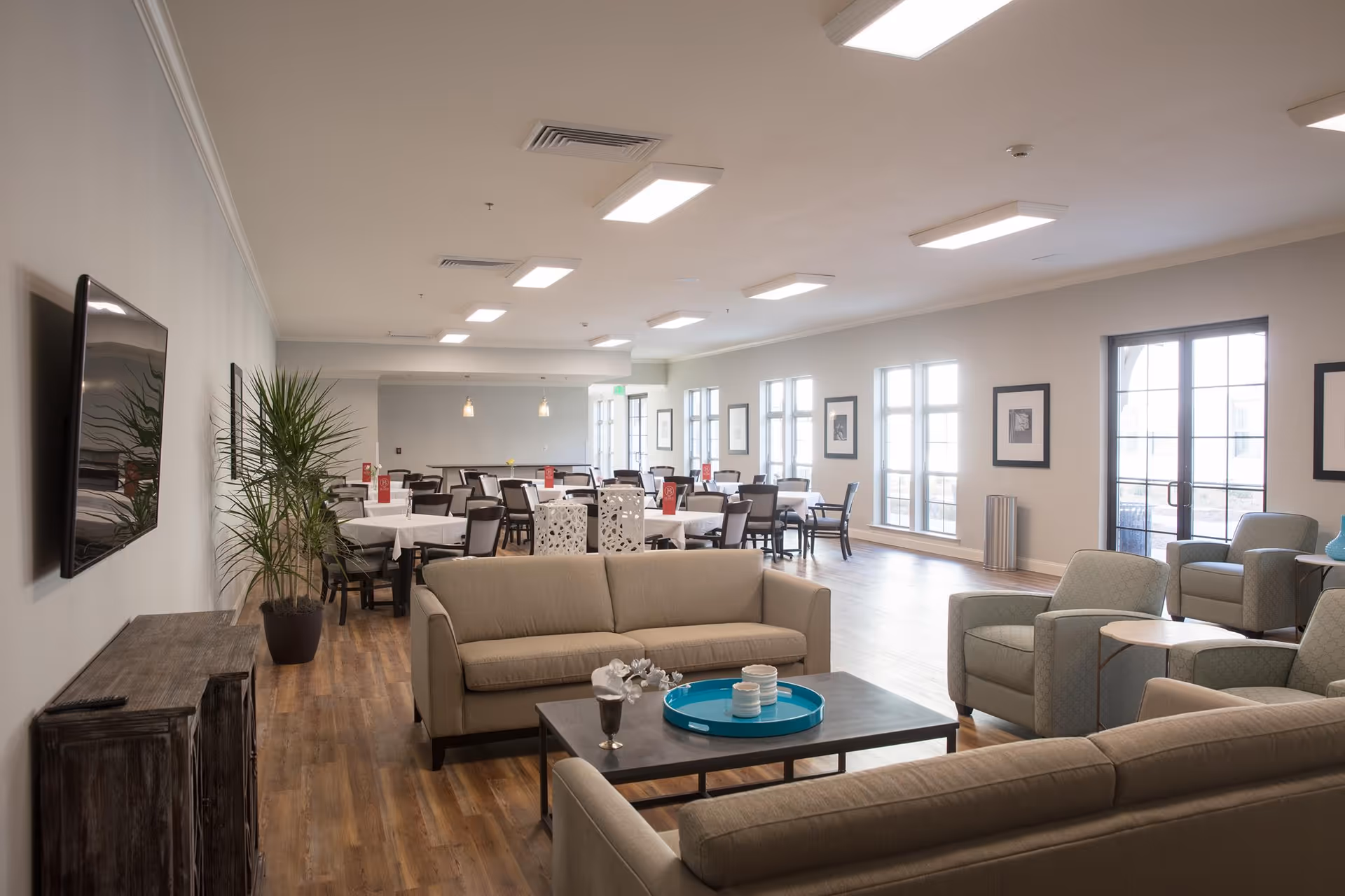 A spacious and well-lit common area in a senior living facility featuring beige sofas and armchairs arranged around a coffee table with decorative items. In the background, there are multiple dining tables with chairs covered in white tablecloths, and large windows allowing natural light to fill the room. A flat-screen TV is mounted on the wall above a wooden cabinet, and a tall potted plant is placed nearby.