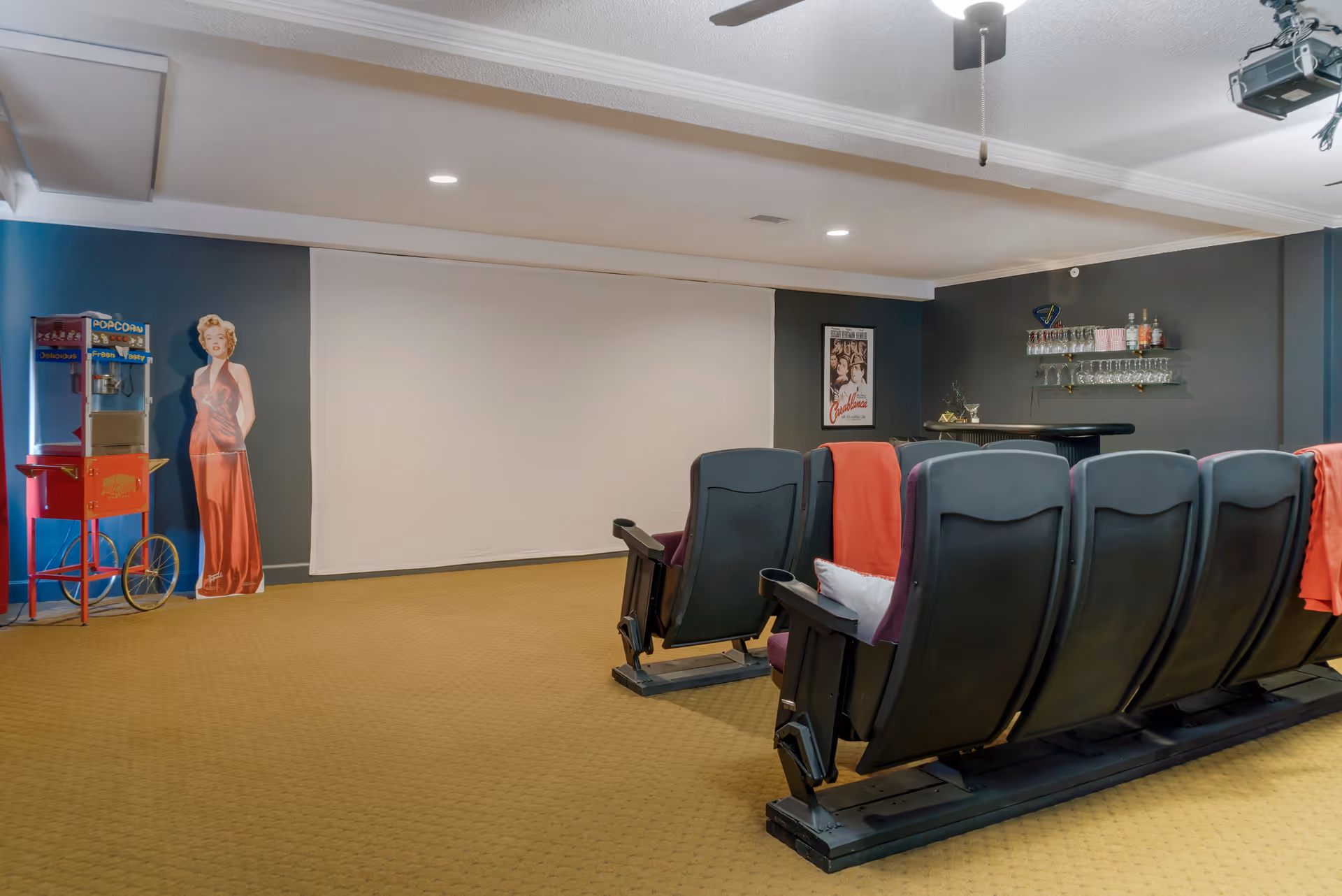 A small home theater room with a row of theater-style seats facing a large blank projector screen. There is a popcorn machine and a life-size cutout of a woman in a red dress on the left side. On the right side, there is a small bar area with shelves holding glasses and bottles. The walls are painted dark gray and the floor is carpeted in a light brown color.