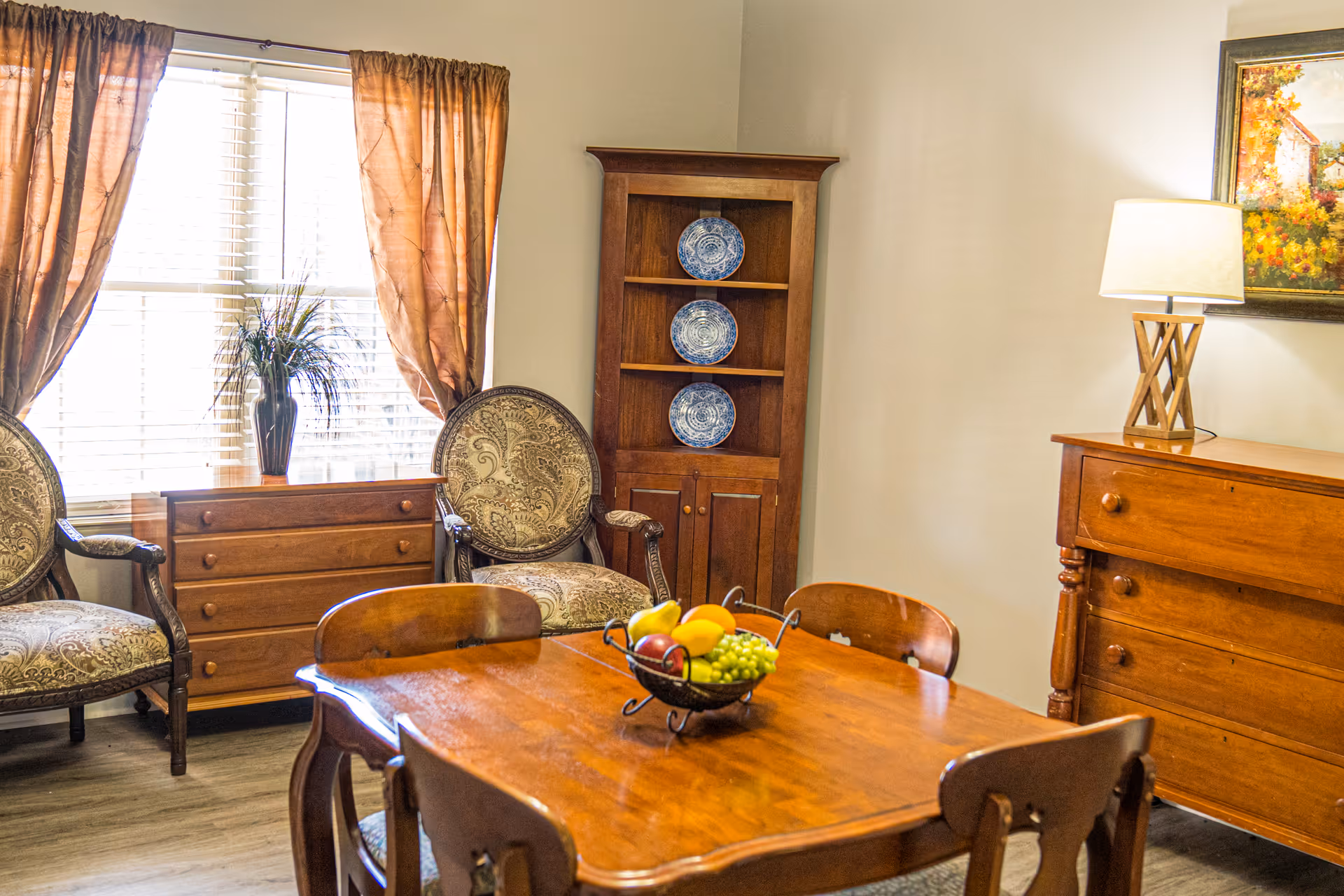 A cozy dining area with a wooden table and four chairs. On the table is a fruit bowl containing bananas, grapes, and apples. Behind the table are two upholstered armchairs with patterned fabric, a wooden dresser, and a tall wooden cabinet displaying three decorative blue and white plates. A window with brown curtains lets in natural light, and a wooden sideboard with a lamp and a framed painting is visible on the right wall.