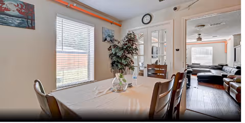 Interior view of a dining area in Agape Assisted Living featuring a wooden dining table with four chairs, a tablecloth, a glass bowl with a plant, and a water bottle. There is a large window with blinds on the left wall, two paintings hanging on the walls, and a clock above a set of French doors leading to another room with a black sectional sofa and additional seating.