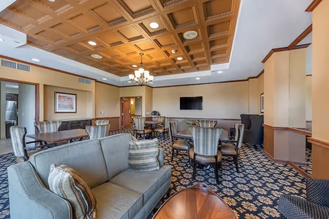 A spacious living room area in a senior living facility with patterned carpet, a wooden coffered ceiling, a chandelier, multiple seating arrangements including sofas and chairs, a wall-mounted flat screen TV, and framed artwork on the walls.