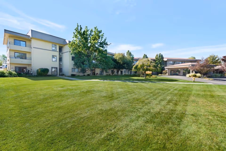 Wide green lawn in front of a multi-story senior living building with trees and a covered entrance under a blue sky.