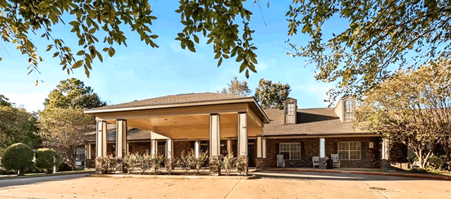 Single-story brick senior living building with a covered entrance portico supported by columns, driveway, and surrounding trees under a blue sky.