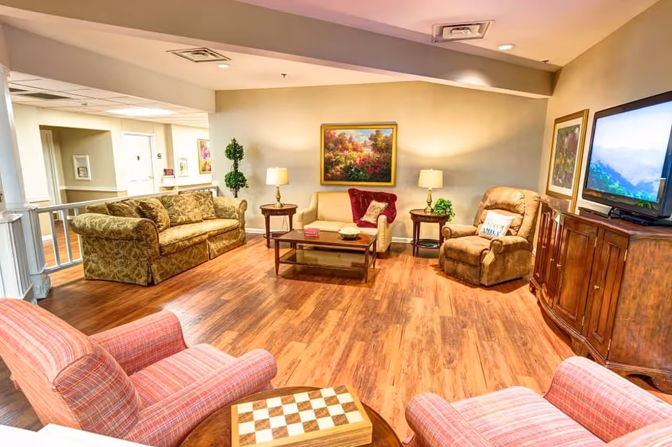 A cozy living room in an assisted living community featuring a variety of seating options including a patterned sofa, a beige loveseat, a brown recliner, and two striped armchairs. The room has wooden flooring, a wooden coffee table, side tables with lamps, framed artwork on the walls, and a flat-screen TV on a wooden cabinet.