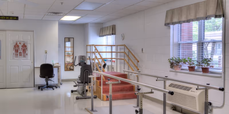 A rehabilitation room in Carlyle Healthcare Center featuring parallel bars for walking exercises, a small set of carpeted stairs with handrails, a stationary exercise bike, a chair and desk, anatomical posters on the door, two windows with potted plants on the windowsill, and a wall-mounted air conditioning unit.