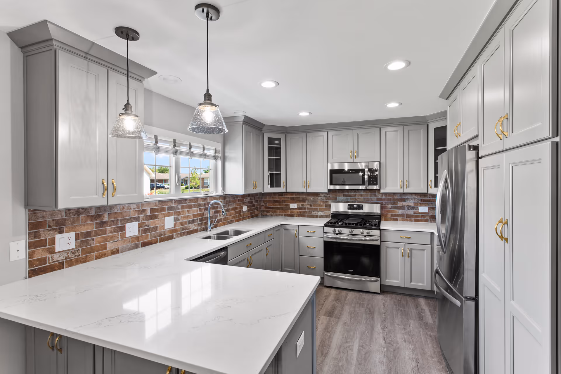 Modern kitchen with gray cabinets, white marble countertops, stainless steel appliances including a refrigerator, oven, and microwave. The backsplash is made of brown brick tiles. Two pendant lights hang over the countertop, and there is a window above the sink letting in natural light.