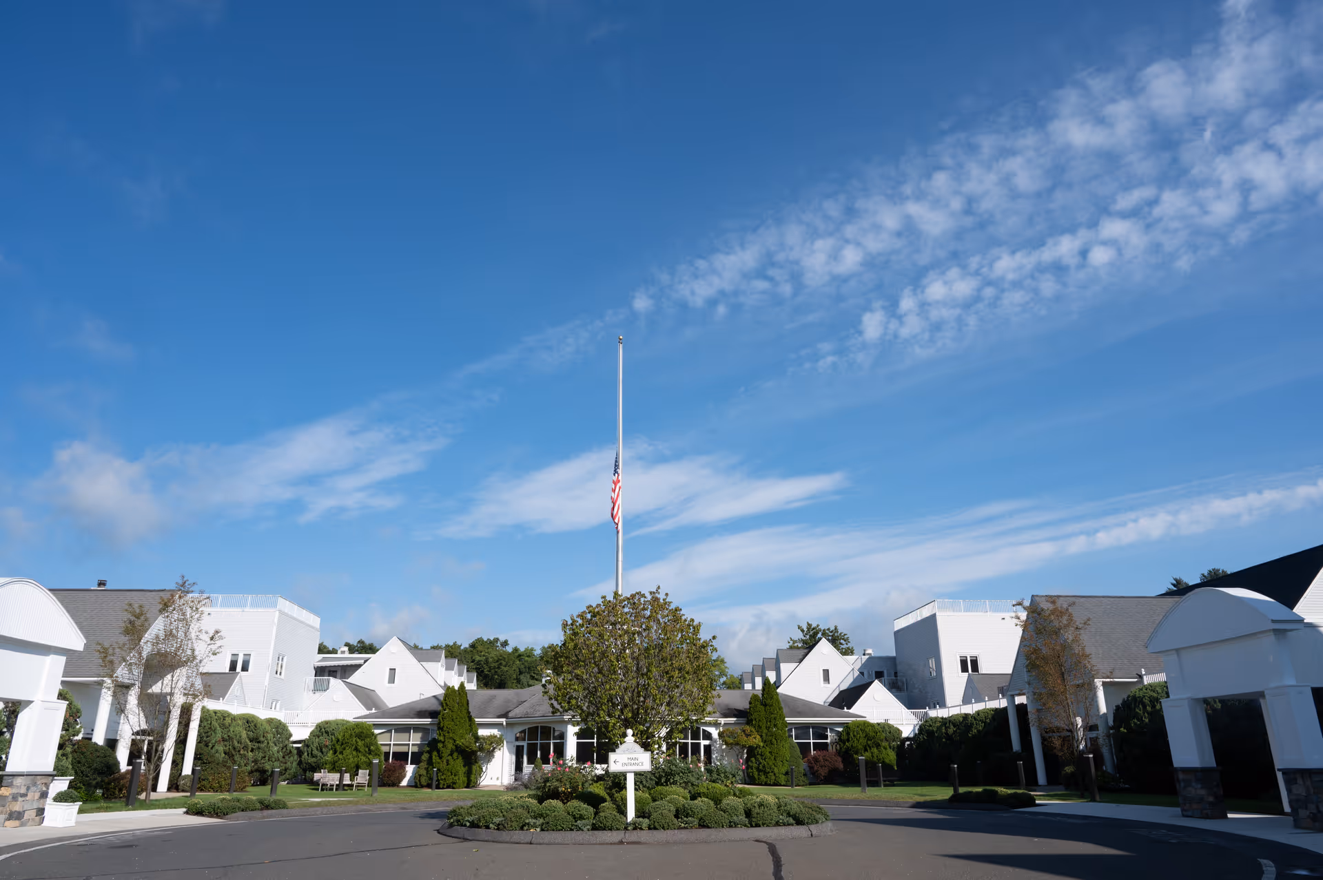 Exterior view of Pomperaug Woods senior living facility showing a circular driveway with a landscaped island in the center featuring a flagpole with an American flag at half-mast. The building is white with multiple peaked roofs and surrounded by greenery under a blue sky with scattered clouds.