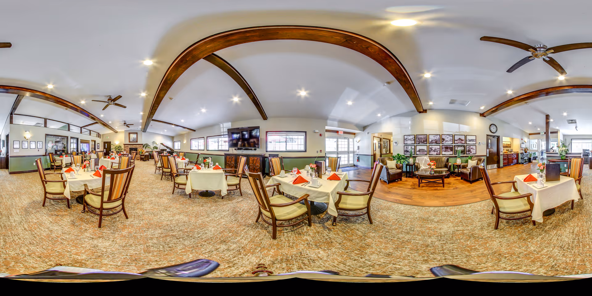 A spacious dining room in a senior living facility with multiple tables covered in white tablecloths, each set with red napkins and glassware. The room features wooden beams on the ceiling, ceiling fans, a carpeted floor, and a seating area with armchairs and a coffee table near the entrance. Large windows allow natural light to fill the space.