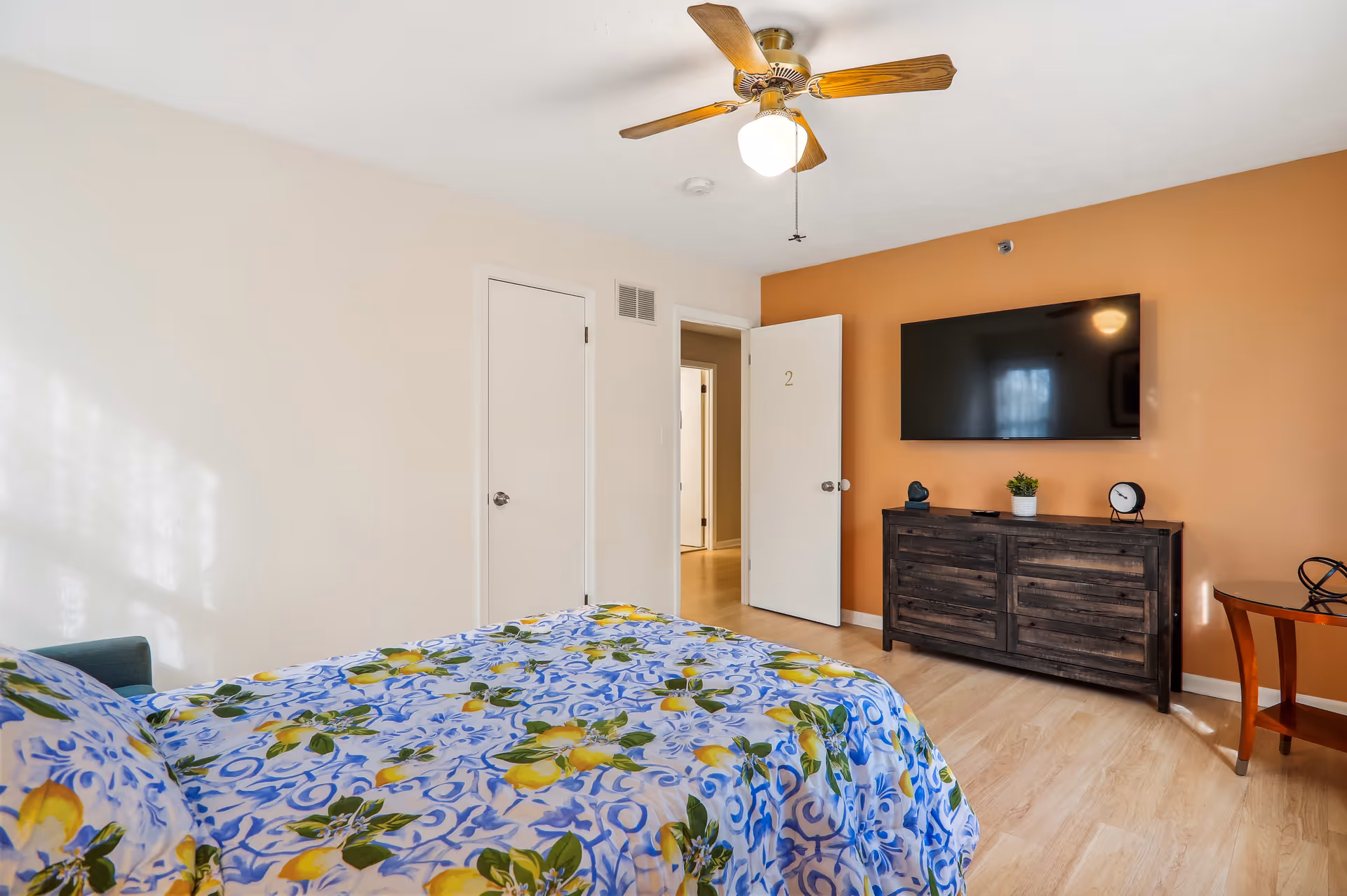 A bedroom with a bed covered in a colorful floral and lemon patterned bedspread. The room has light-colored walls with one accent wall painted orange. A ceiling fan with a light is mounted on the ceiling. There is a dark wooden dresser with a small plant, a clock, and a decorative item on top. A flat-screen TV is mounted on the orange wall above the dresser. Two doors are visible, one closed and one open, showing a hallway.