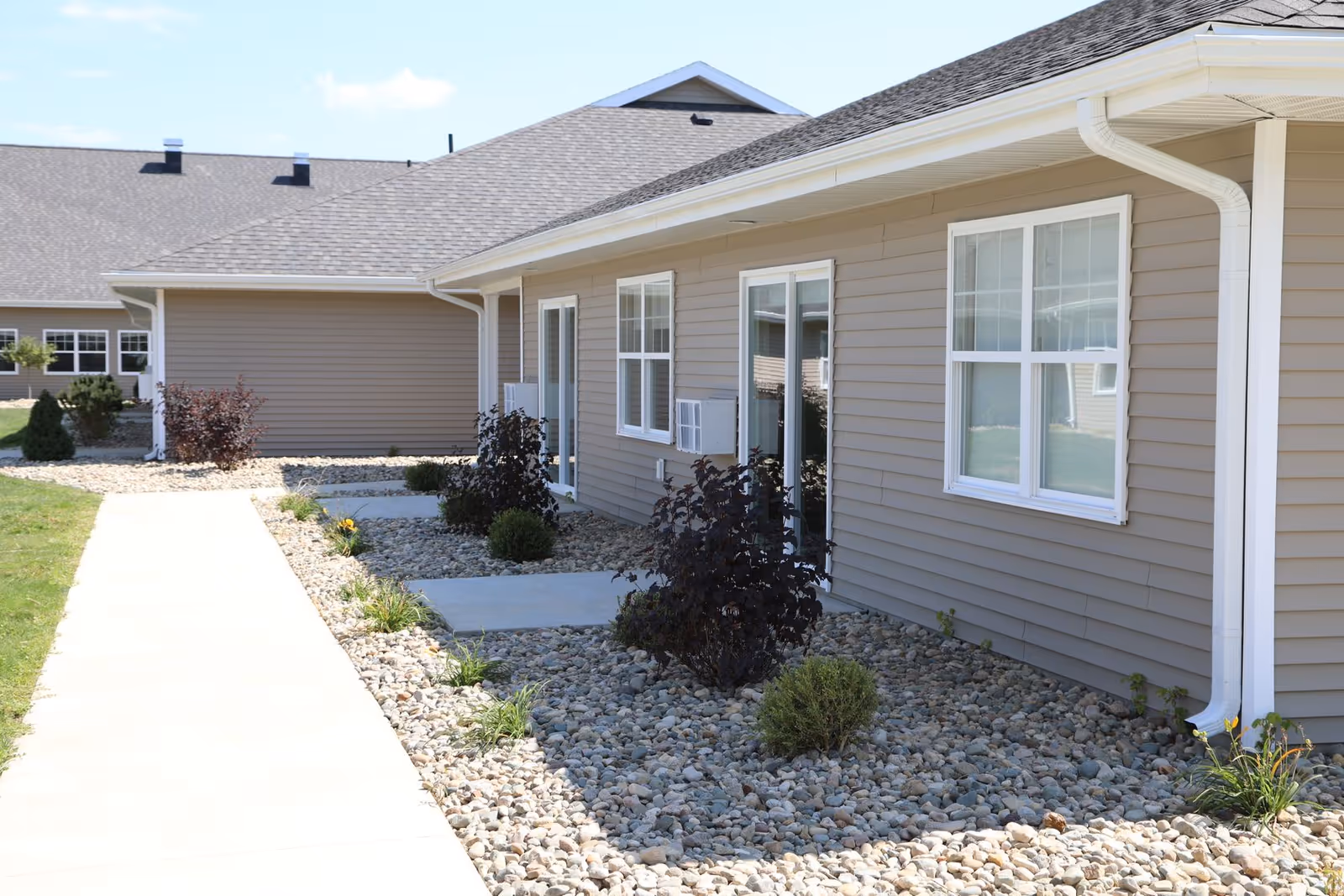Exterior view of a single-story building with beige siding, white-framed windows, and a gray shingled roof. A concrete walkway runs alongside the building, bordered by landscaping with small bushes and rocks. The sky is clear with a few clouds.
