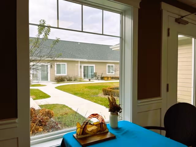 View through a large window showing a courtyard with a walkway, grass, and bushes. Outside, there are single-story buildings with beige siding and green roofs. Inside, a table covered with a blue tablecloth holds a decorative glass sculpture and a small vase with dried flowers. A dark chair is partially visible to the right.