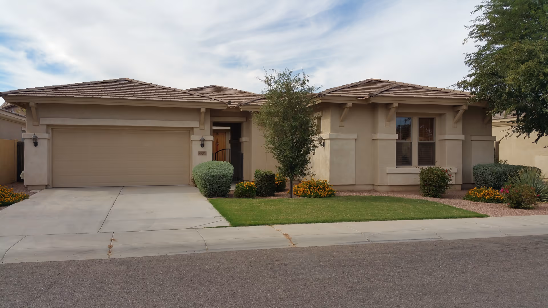 Single-story stucco house with a two-car garage, front lawn and shrubs under a partly cloudy sky.