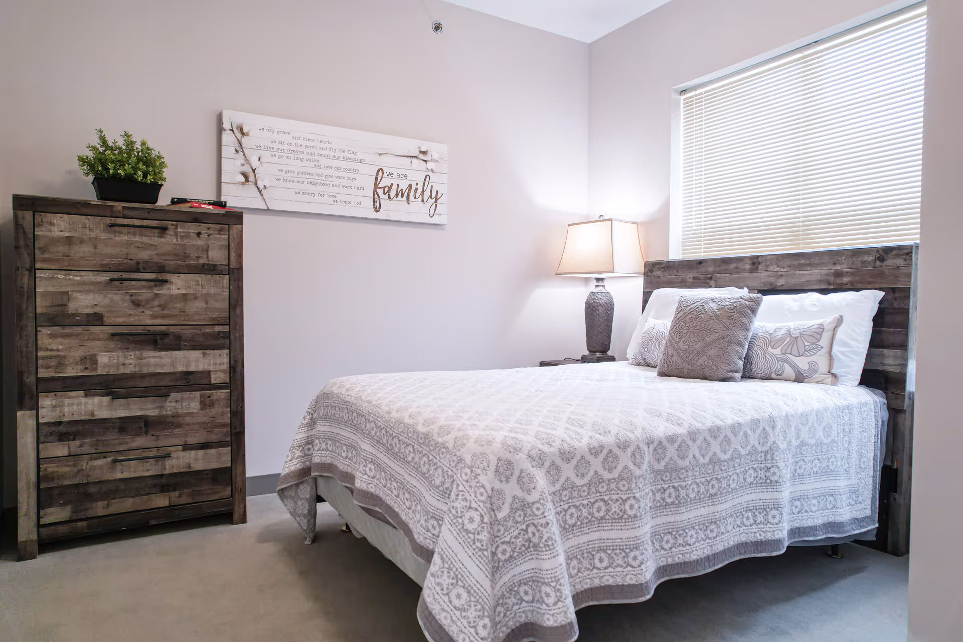 Neatly made bedroom with a wooden headboard and dresser, patterned bedspread, bedside lamp, and wall art that says "family".
