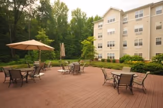 Outdoor patio with tables, chairs, and umbrellas beside a multi-story apartment building surrounded by trees.