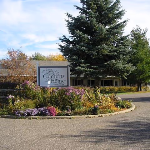 Landscaped circular driveway with a flowerbed and a sign for 'Comforts of Home' in front of a single-story assisted living building and a large evergreen tree.