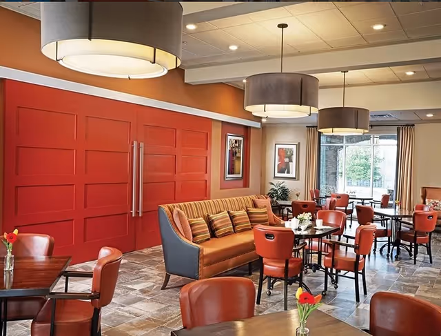 A cozy dining area with multiple round tables and red cushioned chairs. A striped sofa with several pillows is placed against a wall with large red sliding doors. The room has large hanging ceiling lights, framed artwork on the walls, and large windows with beige curtains letting in natural light.