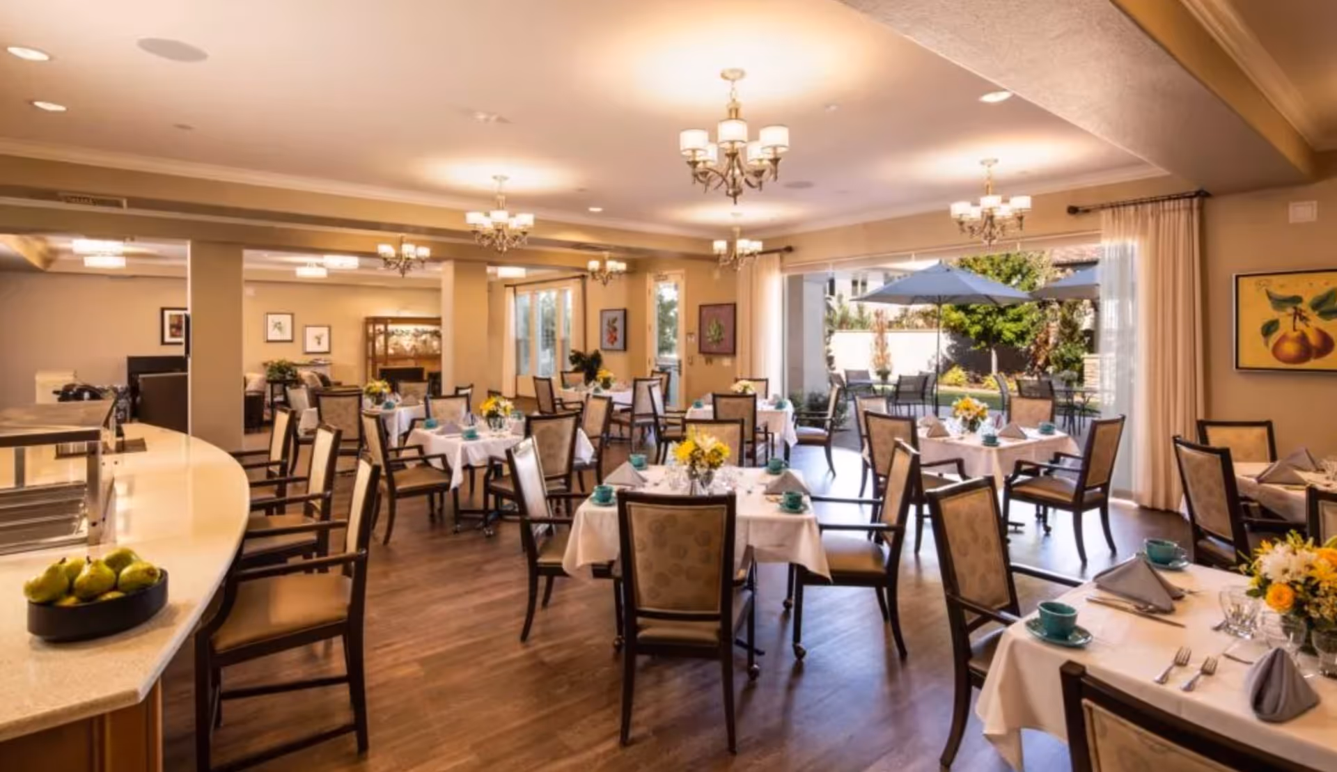 Bright, elegant dining room with multiple tables set with white tablecloths and chairs, chandeliers, and a view to an outdoor patio.