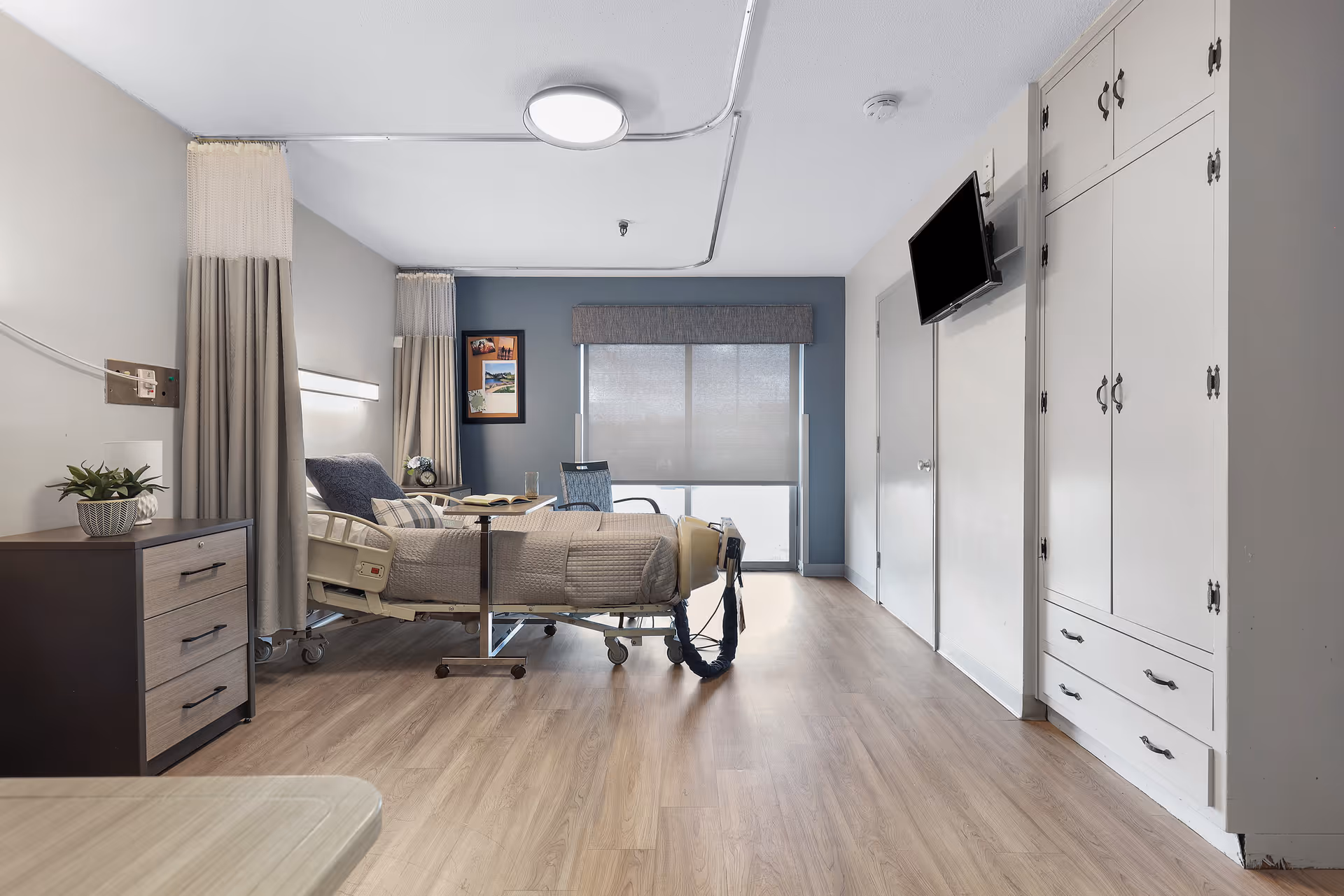 A clean and modern healthcare facility bedroom with a hospital bed, side table with drawers, a chair, a wall-mounted TV, and large windows with blinds. The room has light wood flooring, white walls, and built-in white cabinetry.