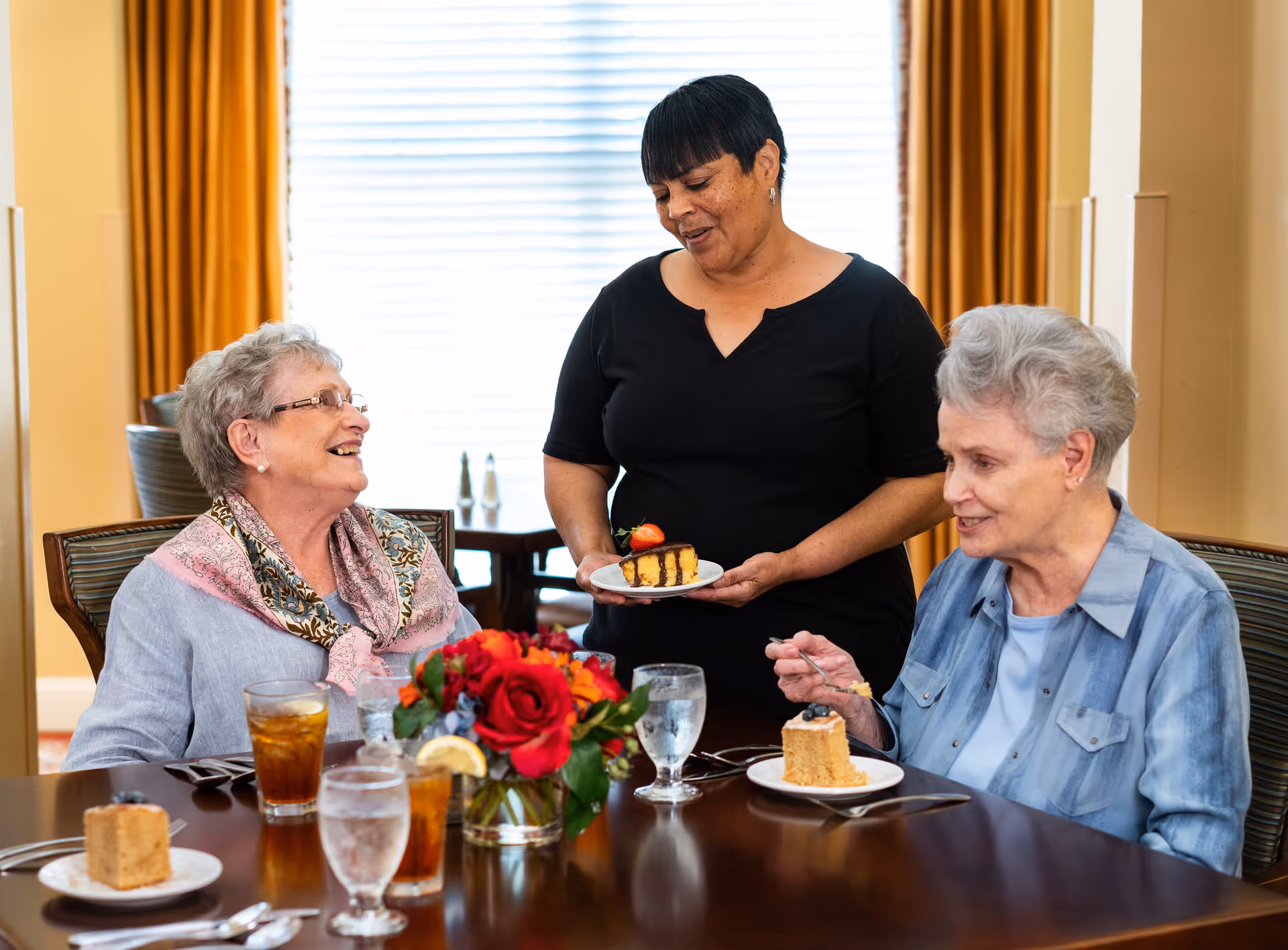 Two elderly women sitting at a dining table with glasses of iced tea and water, each with a slice of cake on a plate. A woman standing beside them is serving a slice of cake. There is a vase with red and orange flowers on the table, and the room has yellow curtains and a window with blinds in the background.