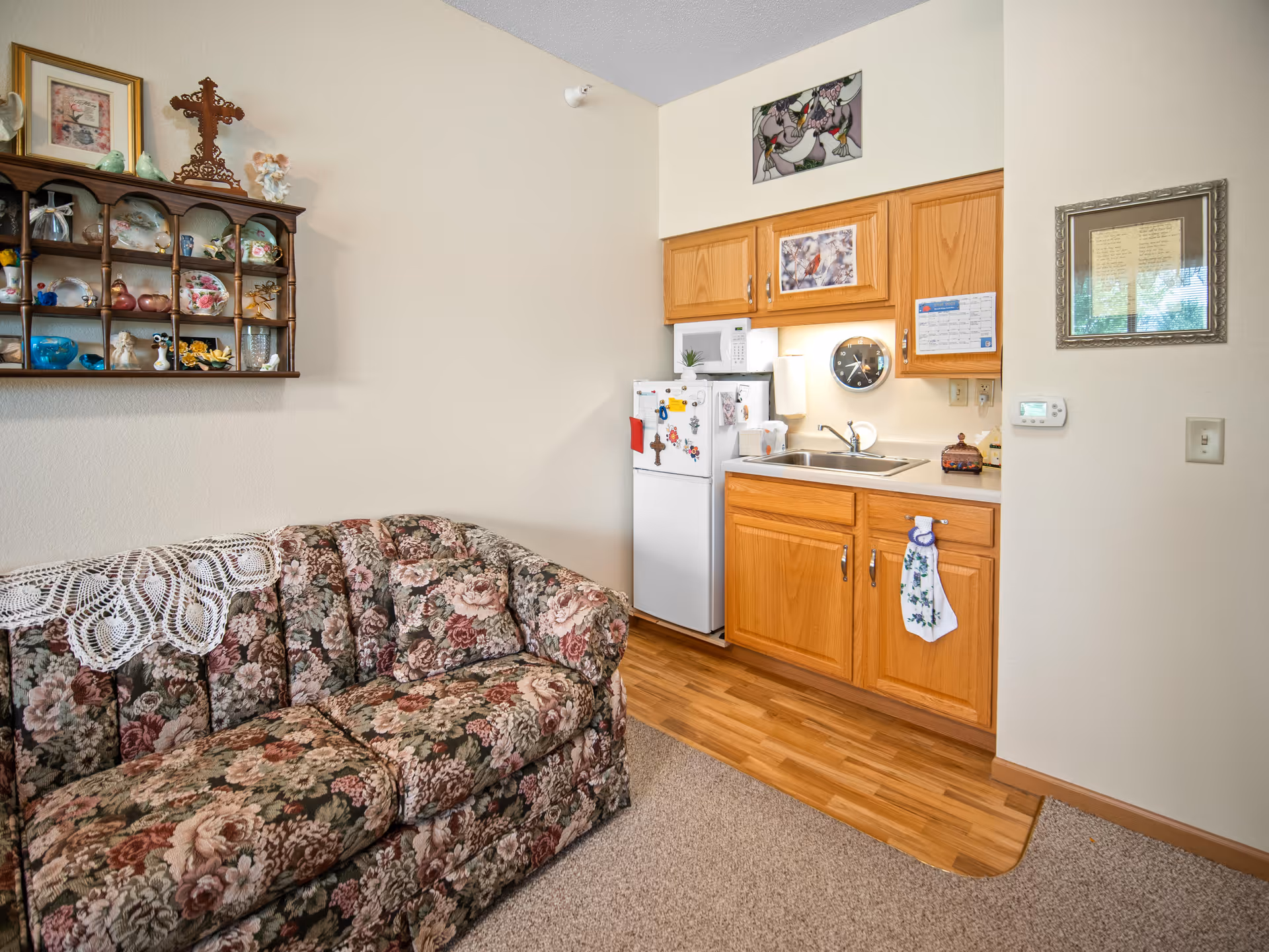 Small living area with a floral upholstered couch, a shelf of knickknacks on the wall, and a kitchenette with oak cabinets and a compact refrigerator.