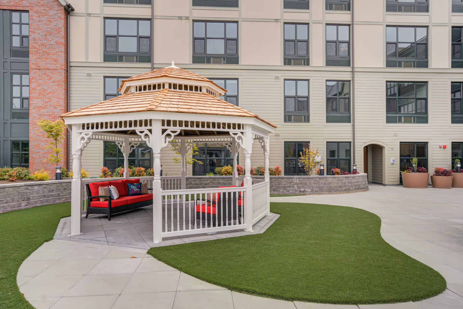 Outdoor gazebo with a wooden roof and white decorative railings, furnished with red cushioned seating and pillows, situated on a paved area with green artificial grass in front of a multi-story building with numerous windows and potted plants.