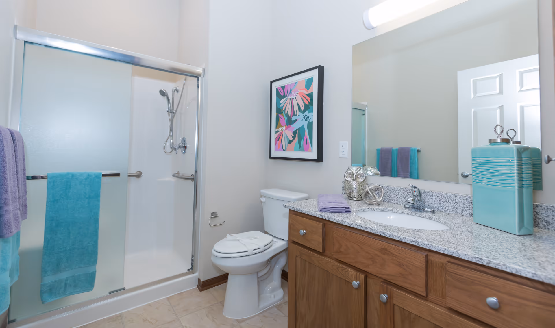 Bright bathroom with a glass-enclosed shower, toilet, and a wooden vanity with granite countertop and mirror.