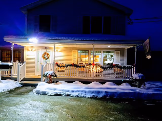 Front exterior view of a two-story house at night with snow on the ground and porch decorated with holiday lights and garlands. The house has a covered porch with white railings and a wreath on the front door. Warm light glows from inside the windows.