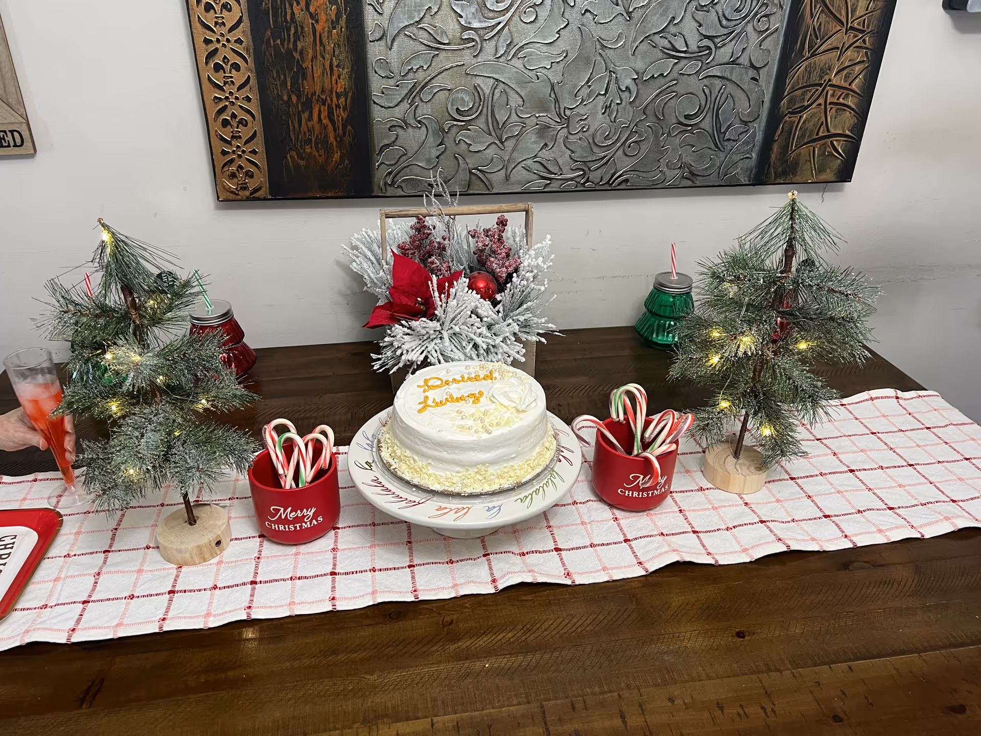 A festive holiday table setting with two small decorated Christmas trees with lights, two red mugs filled with candy canes, a white cake with yellow writing on a decorative plate, and a centerpiece with frosted pine branches and red ornaments. The table has a white and red checkered runner and a wooden surface. A decorative wall art piece is visible in the background.