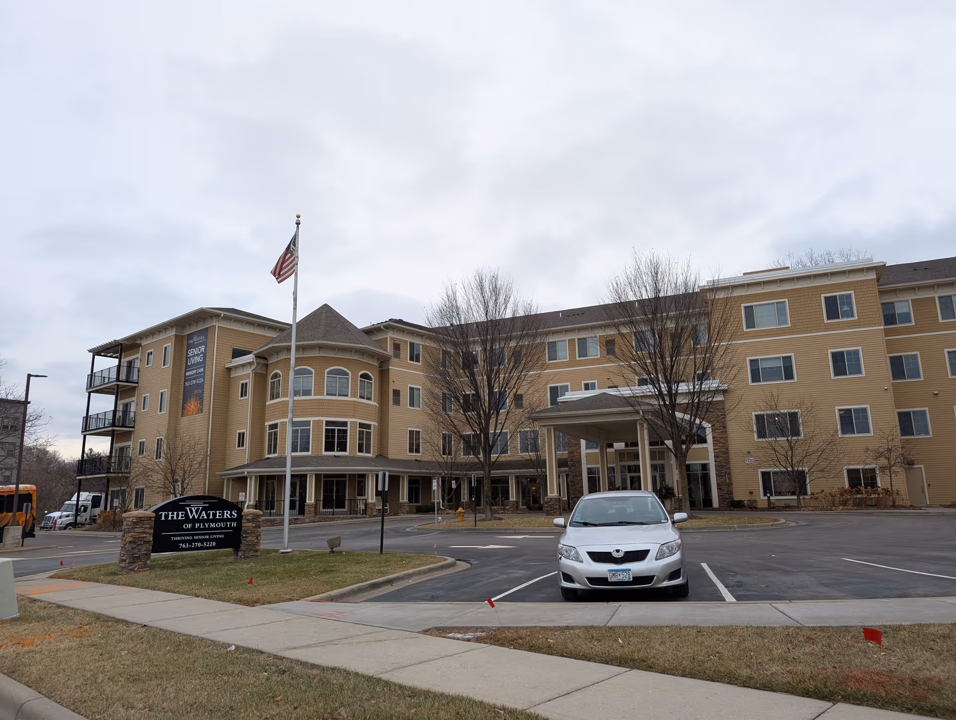 Exterior view of The Waters of Plymouth senior living facility, showing a large multi-story beige building with multiple windows, a covered entrance, an American flag on a flagpole, a sign with the facility name, and a parked silver car in the foreground.