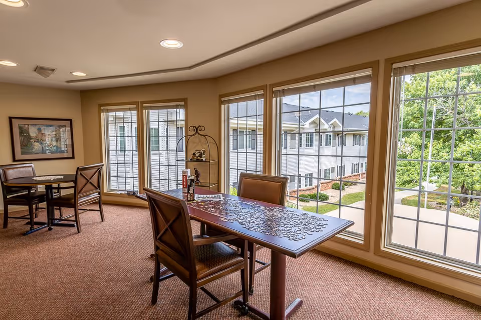 A bright room with large windows overlooking a courtyard and adjacent building. Inside, there are two tables with chairs; one table has a partially completed jigsaw puzzle and puzzle boxes on it. The room has beige walls, carpeted floors, and a framed picture on one wall.