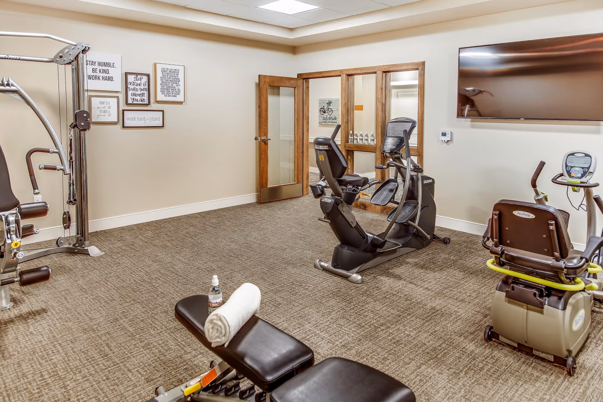 A fitness room with exercise equipment including a recumbent bike, elliptical machine, and a multi-function weight machine. There is a bench with a rolled towel and a water bottle on it. The room has beige walls, carpeted floor, motivational wall art, a large flat-screen TV mounted on the wall, and a wooden door with glass panels.