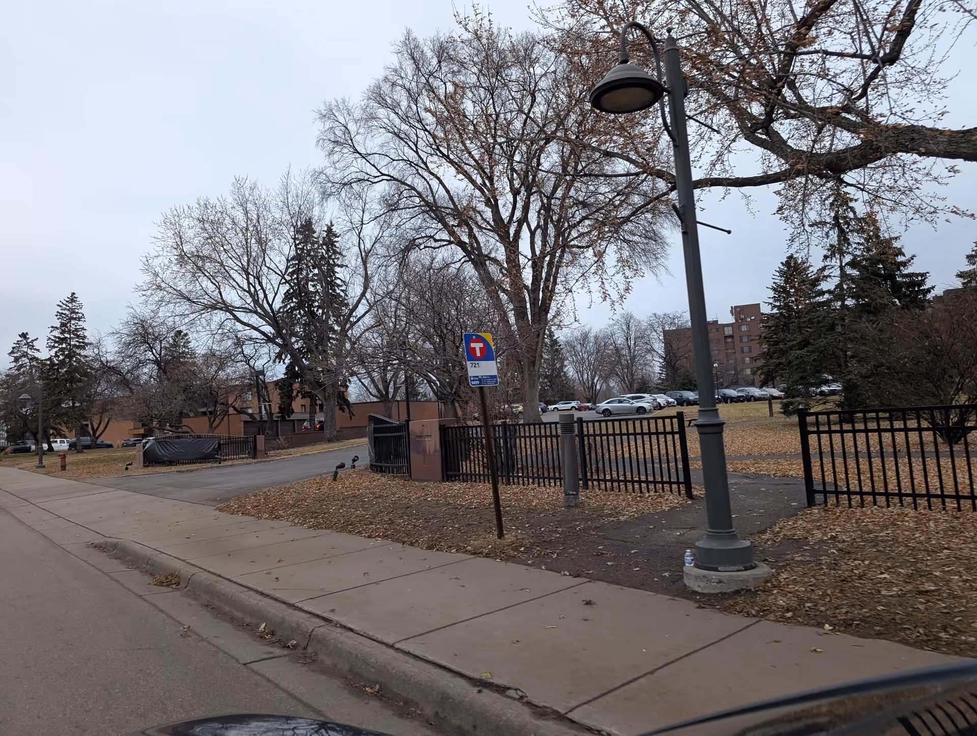 Outdoor view of a senior living facility area with a sidewalk, streetlamp, leafless trees, a black metal fence, parked cars in the background, and a bus stop sign.