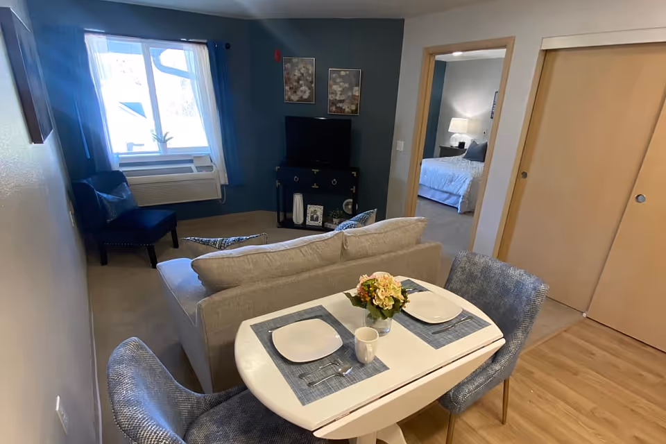 Small furnished living and dining area in a senior apartment with a table set for two, sofa, TV, and a bedroom visible through a doorway.