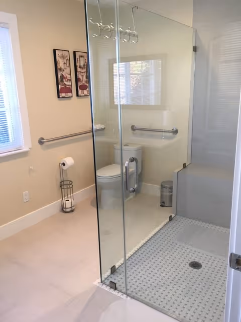 A clean and modern bathroom with a glass-enclosed shower area featuring a tiled floor. Inside the bathroom, there is a white toilet with a grab bar on the wall behind it and another grab bar on the adjacent wall. A toilet paper holder with extra rolls is positioned near the toilet. Two framed pictures hang on the beige wall above the grab bar. A window with blinds allows natural light into the room.