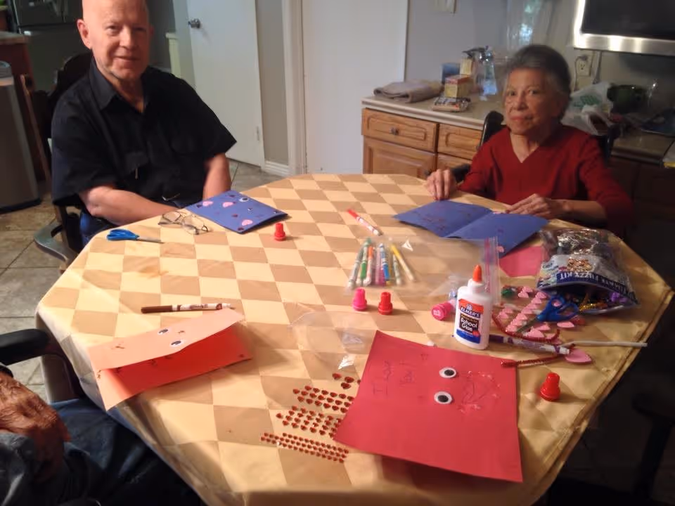 Two elderly individuals sitting at a round table covered with a checkered tablecloth, engaging in arts and crafts activities. The table has various craft supplies including glue, markers, scissors, and decorated paper. The setting appears to be a cozy kitchen or dining area with wooden cabinets in the background.