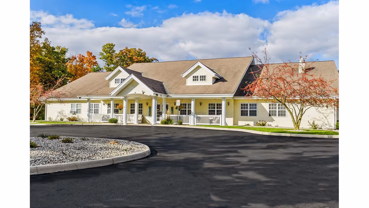 Exterior view of a single-story senior living facility building with a beige facade, white trim, and a covered entrance. The building is surrounded by a paved driveway and landscaped areas with trees showing autumn foliage under a partly cloudy blue sky.