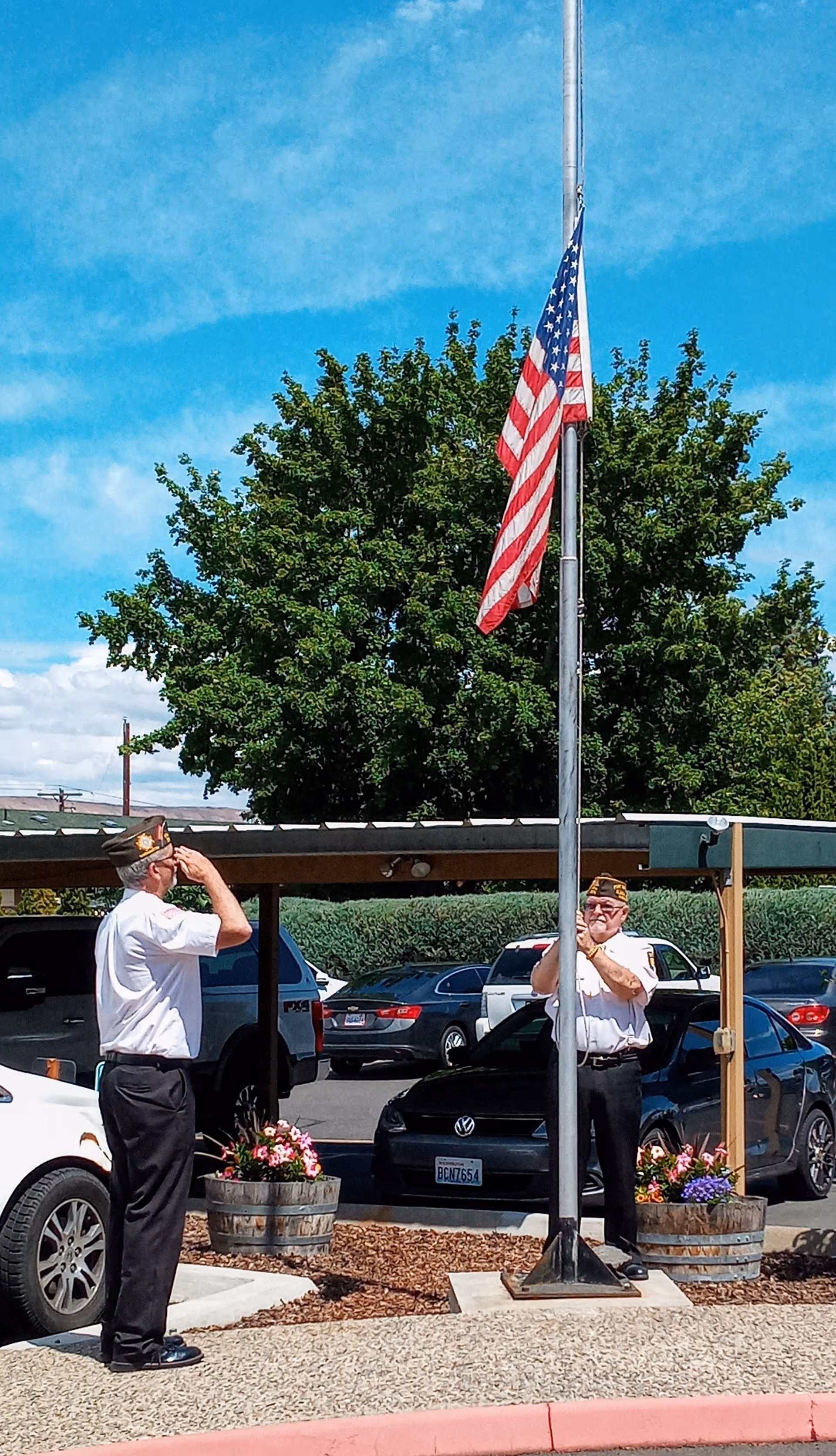 Two elderly men in white shirts and black pants, wearing military hats, are standing outdoors near a flagpole with an American flag at half-mast. One man is saluting the flag while the other is holding the flagpole. There are cars parked under a carport and a large green tree in the background under a blue sky.