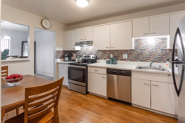 A modern kitchen with white cabinets, a stainless steel oven, dishwasher, and refrigerator. The backsplash features a colorful mosaic tile design. There is a wooden dining table with chairs and a bowl of red apples on it. A clock is mounted on the wall above a pass-through window to an adjacent room.