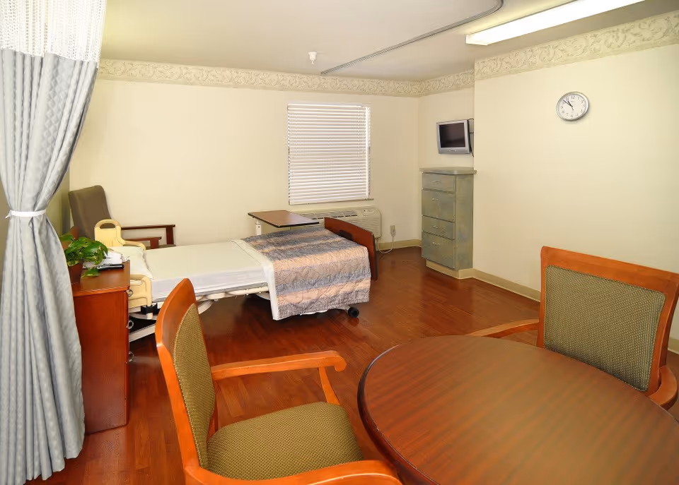 Bright patient bedroom with a hospital-style bed, bedside furniture, a round wooden table and chairs, wall-mounted TV, and hardwood floor.