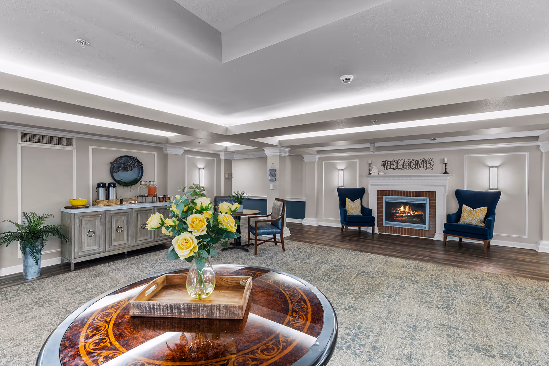A spacious and elegant common area with a fireplace, two blue armchairs with yellow pillows, a round wooden table with a vase of yellow roses, a sideboard with coffee and juice dispensers, and a welcoming sign above the fireplace. The room features soft lighting, neutral walls with white trim, and a patterned carpet.