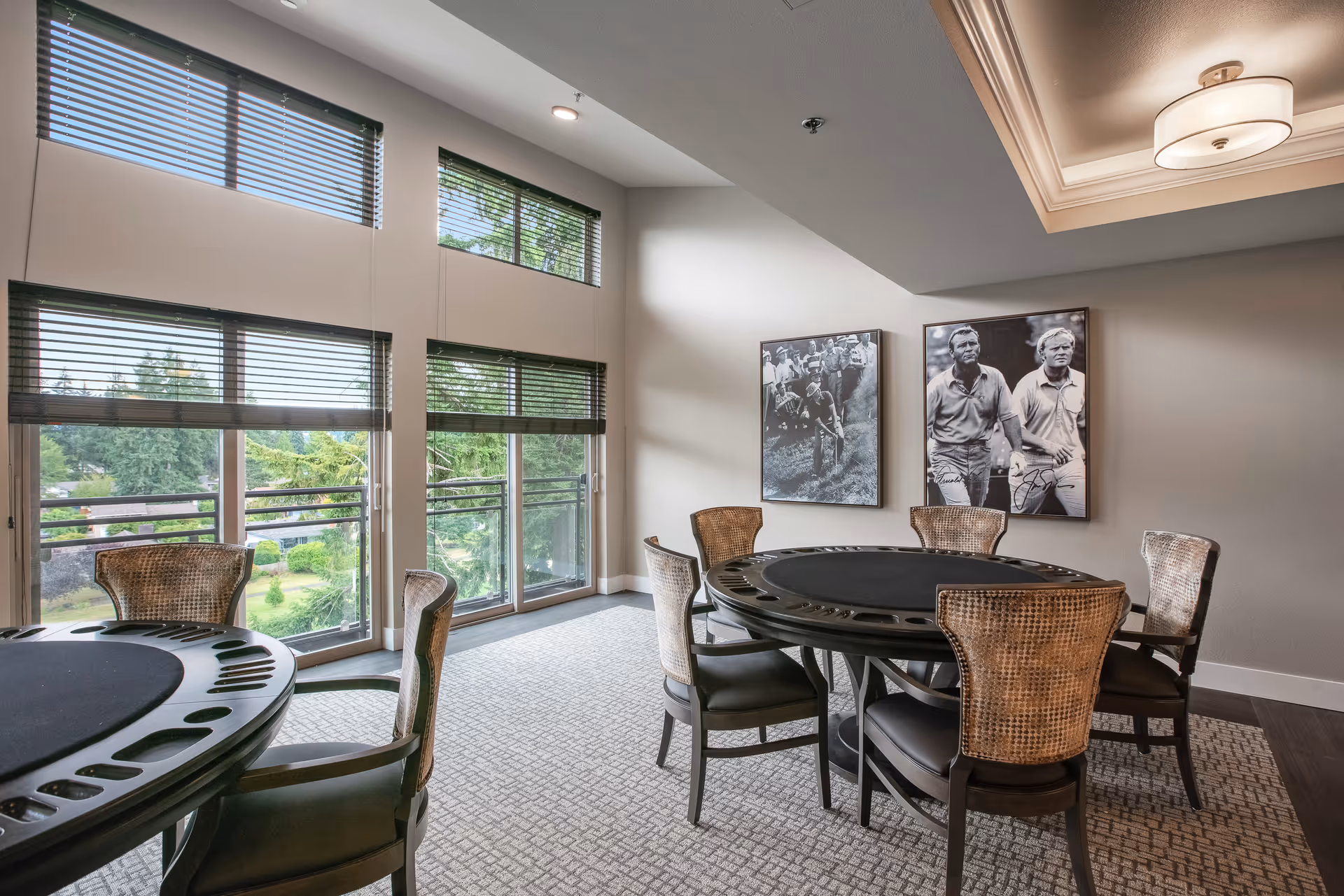 A well-lit room with large windows featuring blinds, overlooking a green outdoor area. The room contains two round poker tables with cup holders and six wicker-backed chairs around one table. Two black and white framed photographs of people are hung on the wall. The ceiling has a recessed light fixture with a modern design.