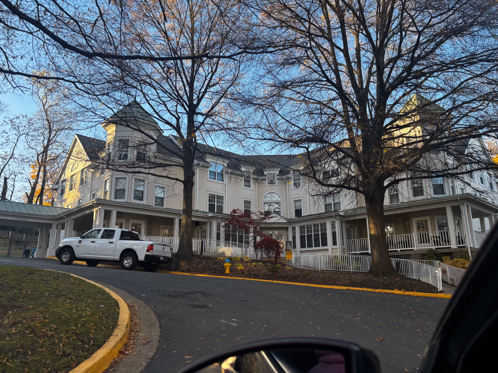 Exterior view of a large, multi-story senior living facility building with beige siding and multiple windows. There are two large leafless trees in front, a white pickup truck parked near the entrance, and a curved driveway with a yellow curb. The sky is clear and blue.