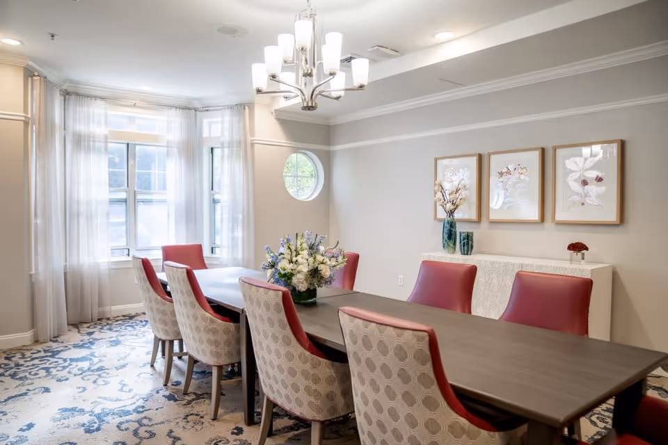 A bright dining room with a long rectangular table surrounded by eight chairs, four with red backs and four with patterned upholstery. The room features large windows with sheer white curtains, a modern chandelier, a patterned carpet, and a sideboard with three framed floral artworks and decorative vases.