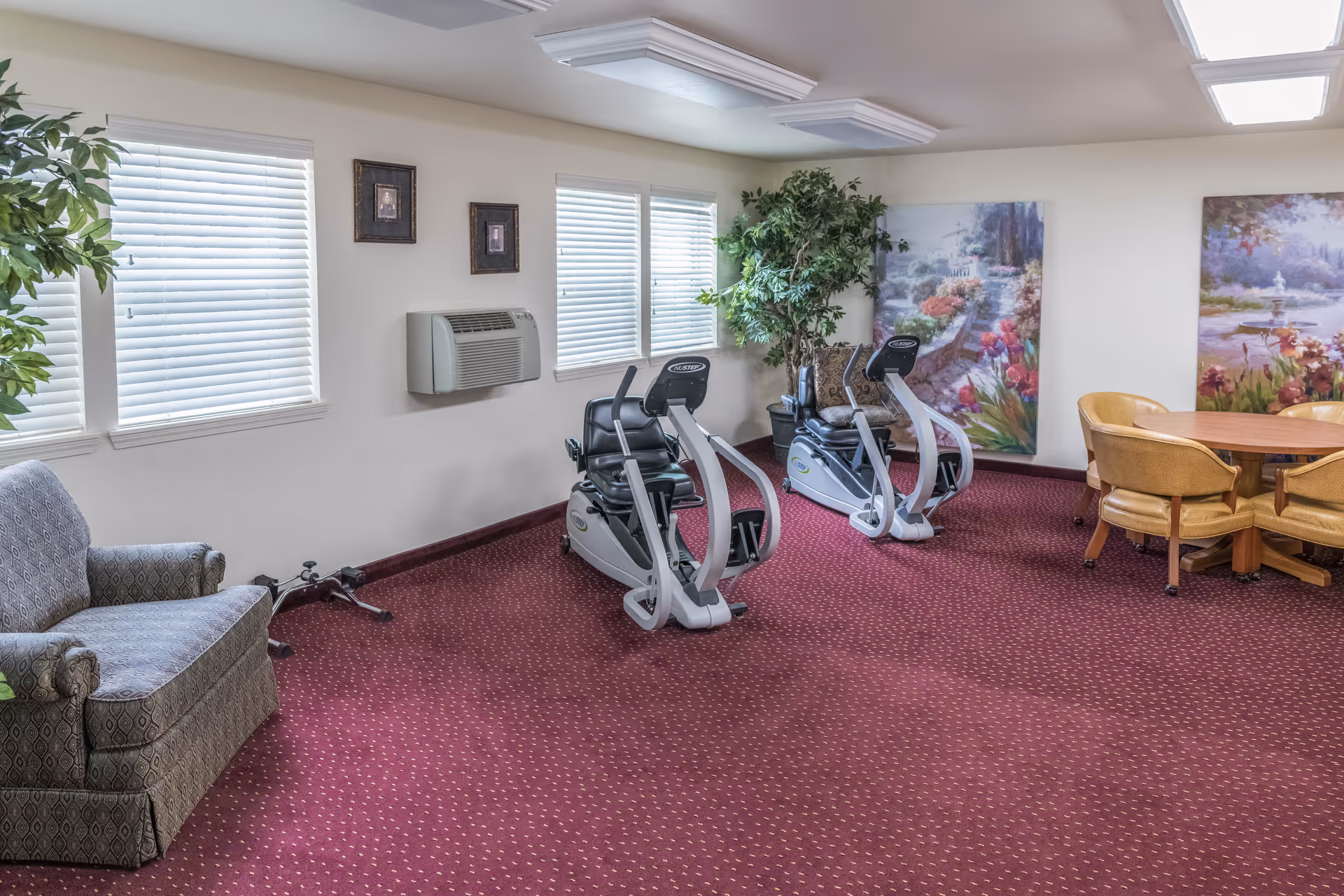 Carpeted common room with two recumbent exercise bikes, upholstered seating, a round table with chairs, and floral wall art.