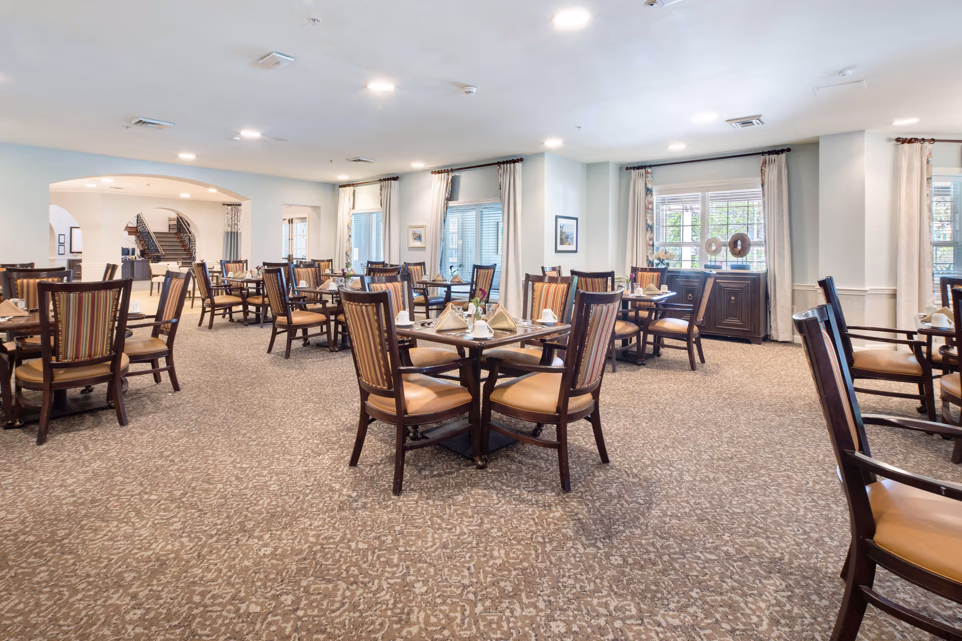 A spacious dining room with multiple wooden tables and chairs arranged neatly. Each table is set with napkins, cups, and small flower vases. The room has large windows with curtains allowing natural light to brighten the space. The carpeted floor and soft lighting create a warm and inviting atmosphere.