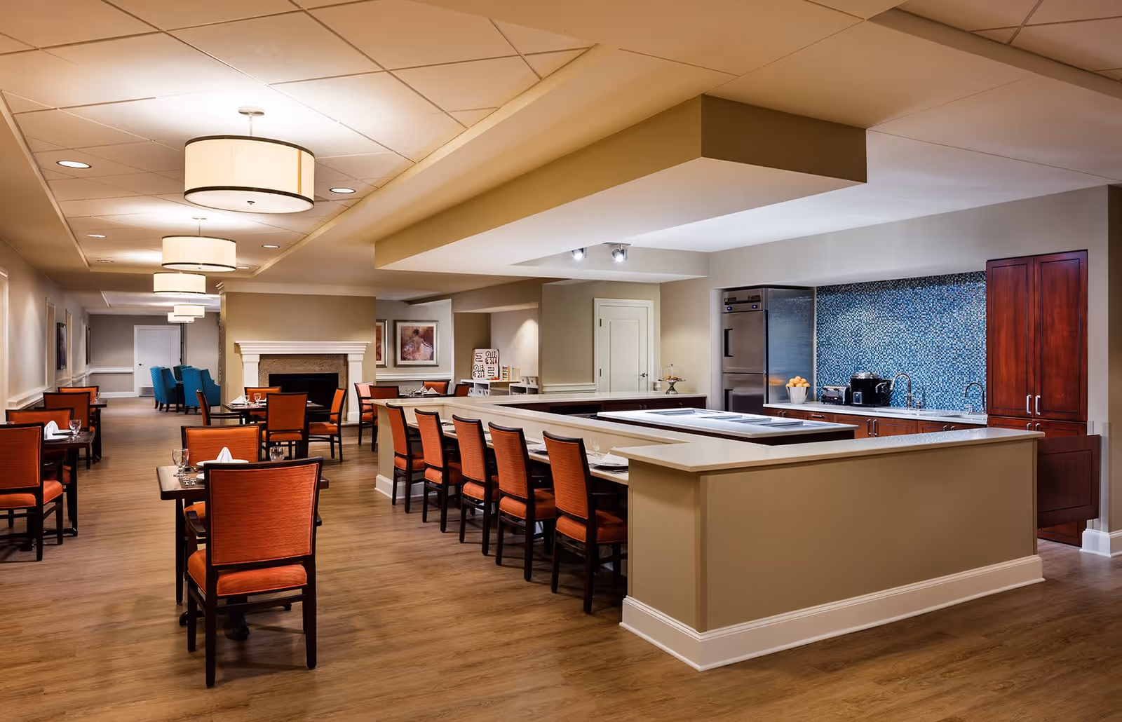 A spacious dining area in a senior living facility featuring multiple tables with orange cushioned chairs, a long counter with additional seating, a kitchen area with stainless steel appliances and a blue mosaic tile backsplash, warm lighting from ceiling fixtures, and a fireplace in the background.