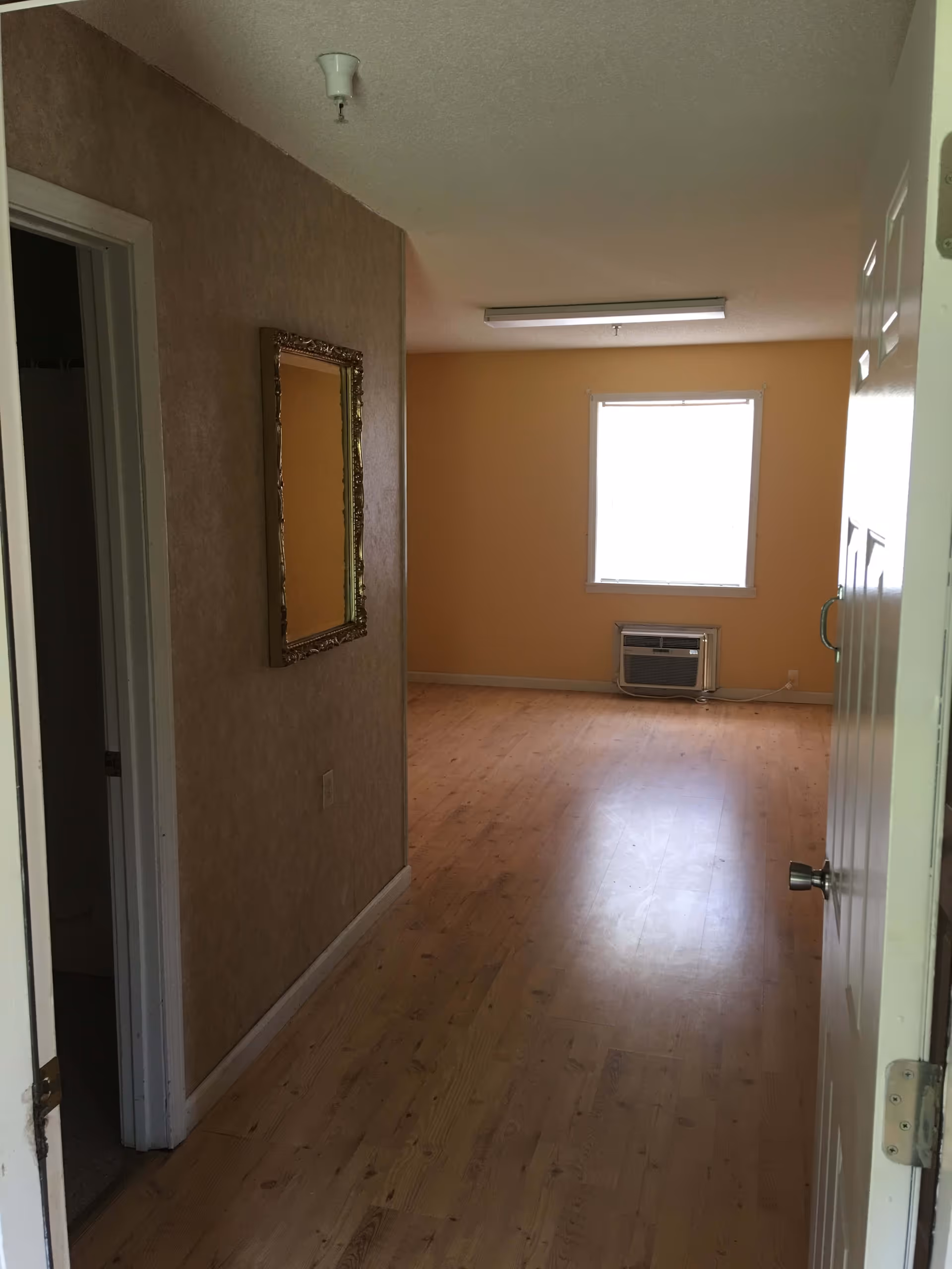 Empty interior hallway leading to a sunlit room with wood floors, a window and a wall air conditioner.