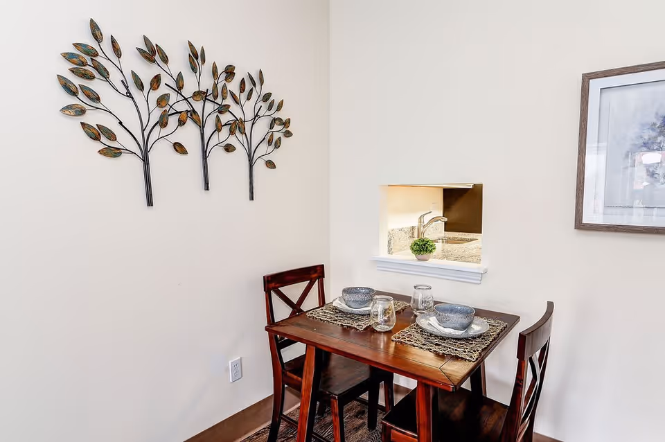 A small dining area with a wooden table set for two with bowls, plates, glasses, and woven placemats. Two wooden chairs are positioned at the table. On the wall above the table is a decorative metal wall art piece shaped like three trees with leaves. There is a small pass-through window to a kitchen area with a granite countertop and a small potted plant. A framed picture hangs on the adjacent wall.